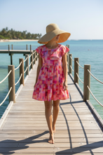A woman walking barefoot on a wooden pier over turquoise water, wearing a pink and purple geometric patterned tiered dress.