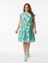 A woman modeling a short-sleeve tiered dress with a colorful geometric print in shades of blue, green, yellow, pink, and white, paired with teal sandals, against a plain white studio background.