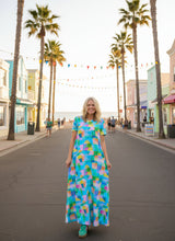 A woman stands on a black and white patterned rug against a light gray wall, wearing a short-sleeved maxi dress with a colorful geometric block pattern in shades of blue, green, pink, yellow, and orange.